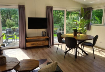 Bright lodge living area with dining table, black chairs, TV, wooden furniture, and garden view outside windows.