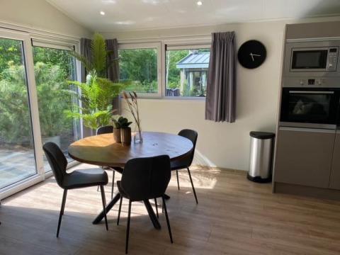 Comedor moderno en una cabaña con mesa redonda, cuatro sillas negras, ventanas grandes y plantas verdes.