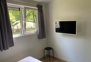 Chambre lumineuse avec fenêtre, télé murale, rideaux gris, tabouret noir et sol en bois dans un lodge.