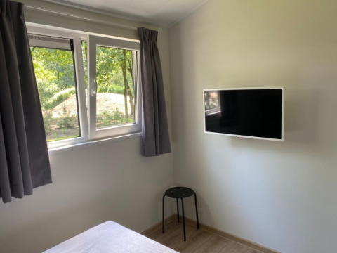 Chambre lumineuse avec fenêtre, télé murale, rideaux gris, tabouret noir et sol en bois dans un lodge.