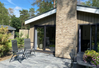 Cabaña moderna con terraza, muebles de jardín y amplios ventanales rodeada de árboles verdes.