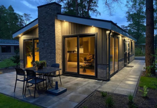 Foto al atardecer de una moderna cabaña con grandes ventanales, terraza y luces exteriores en la naturaleza.