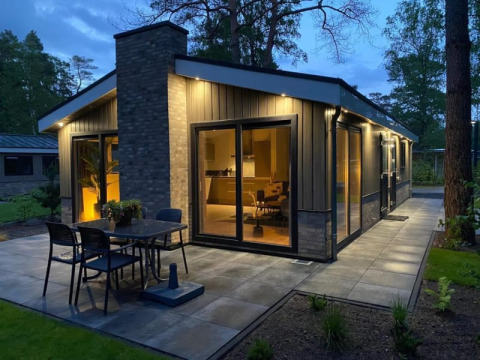 Foto al atardecer de una moderna cabaña con grandes ventanales, terraza y luces exteriores en la naturaleza.