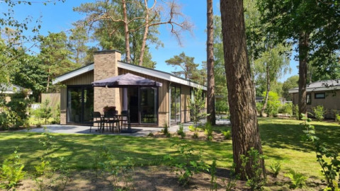 Acogedora cabaña moderna con amplio jardín, rodeada de altos árboles, terraza exterior con mesa y sombrilla.