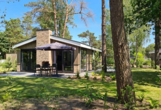 Acogedora cabaña moderna con amplio jardín, rodeada de altos árboles, terraza exterior con mesa y sombrilla.