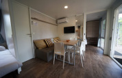 Moderne glampingaccommodatie met sofa, eettafel, wand-tv en houtlook laminaatvloer.