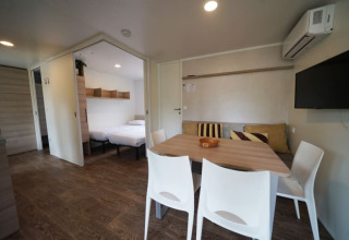 Hébergement glamping moderne avec table à manger, chaises blanches, canapé, TV et chambre confortable.