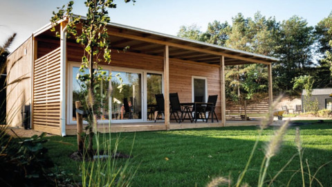 Modernes Holzhaus mit großer Terrasse und Gartenmöbeln, umgeben von Rasen und Bäumen im Sonnenlicht.