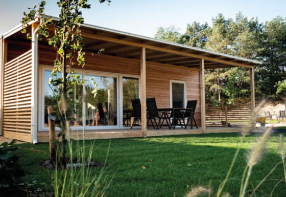 Modernes Holzhaus mit großer Terrasse und Gartenmöbeln, umgeben von Rasen und Bäumen im Sonnenlicht.