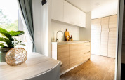 Moderne lodgekeuken met lichte houtkleurige kasten, plant op tafel en groot raam met gordijnen.