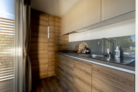 Moderne lodgekeuken met houten kasten, grijs werkblad en groot raam waar zonlicht binnenvalt.