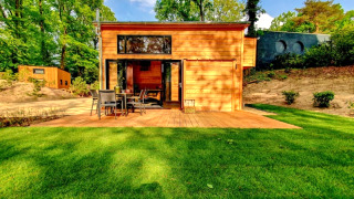 Tiny wooden house with outdoor patio, garden furniture, lush green lawn, and surrounded by tall trees