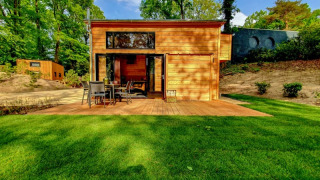 Pequeña casa de madera con terraza, muebles de jardín y césped verde, rodeada de árboles altos