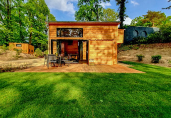 Piccola casa in legno con terrazza, mobili da giardino, prato verde e circondata da alberi alti