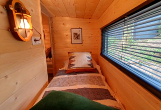 Cozy tiny house bedroom with wood paneling, single bed, and a large window with blinds and forest view.
