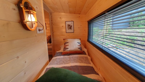 Cozy tiny house bedroom with wood paneling, single bed, and a large window with blinds and forest view.