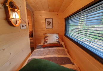 Chambre douillette de tiny house avec murs en bois, lit simple et grande fenêtre avec des stores.