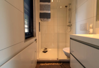 Small, modern bathroom in a tiny house featuring a glass shower, window with blinds, toilet and sink unit.