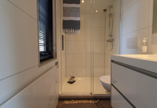 Lille, moderne badeværelse i et tiny house med brusekabine, vindue, toilet og hvidt skabsarrangement.