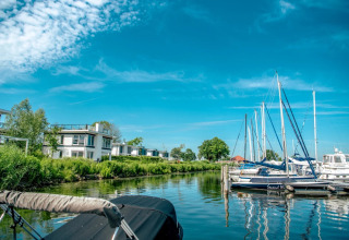 Vakantiepark met moderne glampingwoningen aan het water en aangemeerde zeilboten onder een blauwe lucht.