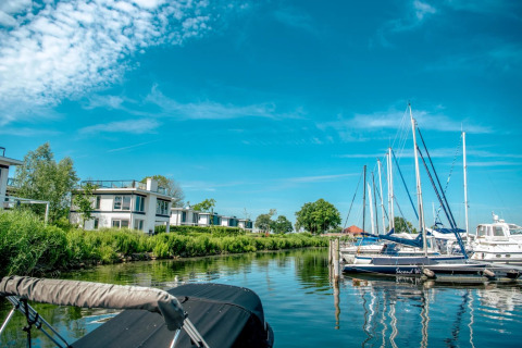 Vakantiepark met moderne glampingwoningen aan het water en aangemeerde zeilboten onder een blauwe lucht.