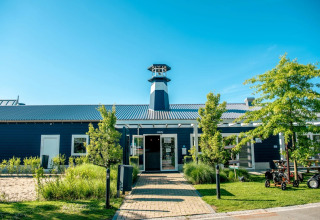 Ingang van een vakantiepark met glamping, blauwe gevel, groene bomen en blauwe lucht op een zonnige dag.