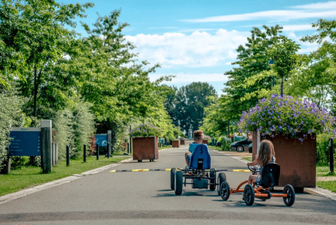 Kinderen rijden op trapkarts langs bomen en bloemen in een vakantiepark met glamping accommodaties.