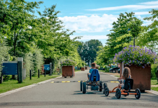 Kinderen rijden op trapkarts langs bomen en bloemen in een vakantiepark met glamping accommodaties.