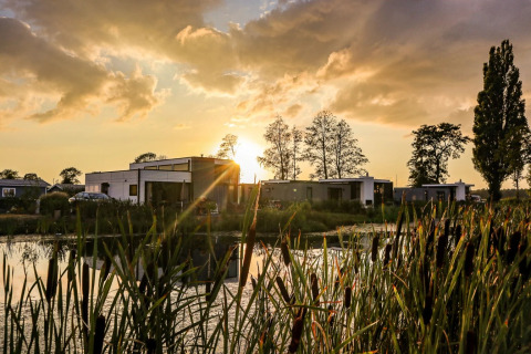 Zonsondergang boven een vakantiepark met glamping accommodaties aan het water, omringd door riet en bomen.