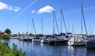 Jachthaven met aangemeerde zeilboten en motorboten onder een blauwe lucht bij een vakantiepark met glamping.