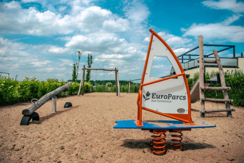 Speeltuin met schommel, wip en veerwip bij EuroParcs Bad Hulckesteijn glamping onder een blauwe lucht.