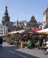 Café al aire libre con personas bajo sombrillas, edificios históricos y torre de iglesia en una plaza soleada.