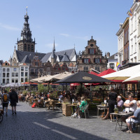 Café al aire libre con personas bajo sombrillas, edificios históricos y torre de iglesia en una plaza soleada.