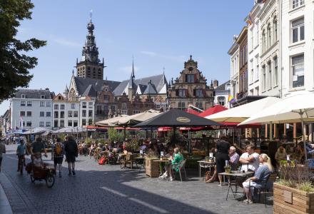Café al aire libre con personas bajo sombrillas, edificios históricos y torre de iglesia en una plaza soleada.