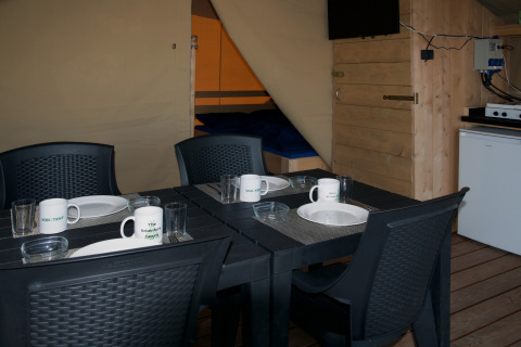 Interior of a safari tent with a black dining table, dishes, mugs, four chairs, fridge, and bed.