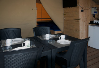 Binnenkant safaritent met zwart eettafel, vier stoelen, servies, mokken, koelkast en bed.