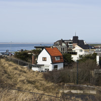 Vista de un parque vacacional con alojamientos glamping, dunas, casas y un faro rojo junto al mar.