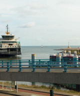 Un ferry entra al puerto, cerca de un parque vacacional con glamping, puente y costa al fondo.