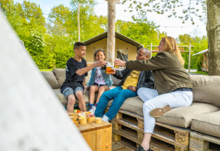 Gezin geniet samen van drankjes en hapjes op een buitenlounge bij een glampingaccommodatie in het vakantiepark.