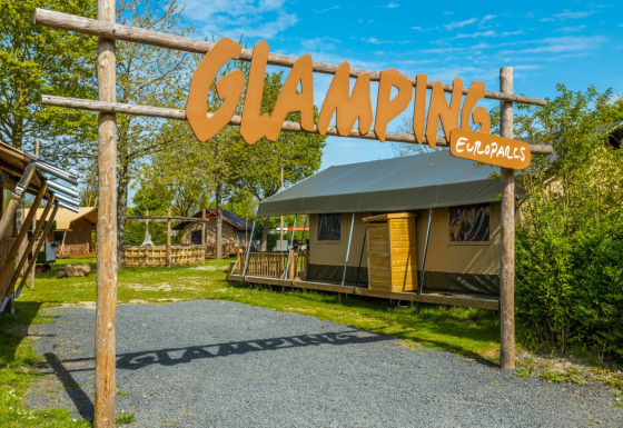 Ingang van een glampingterrein bij een vakantiepark, met luxe tenten en een groot welkomstbord in de lente.