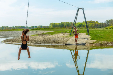 Kind op een kabelbaan boven het water, met een jongen die wacht bij een meer in een vakantiepark.
