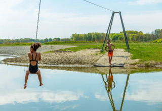Kind op een kabelbaan boven het water, met een jongen die wacht bij een meer in een vakantiepark.