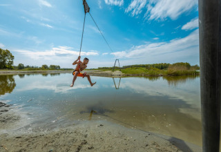 Kind zwaait aan touw boven ondiep water bij zonnige vakantiepark, ideaal voor glamping in de natuur.