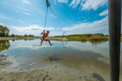 Kind zwaait aan touw boven ondiep water bij zonnige vakantiepark, ideaal voor glamping in de natuur.