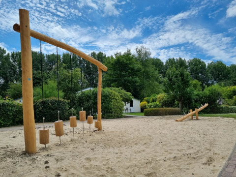 Speeltoestel en wip in een zandbak op een vakantiepark met groene bomen en blauwe lucht op de achtergrond.