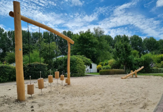 Speeltoestel en wip in een zandbak op een vakantiepark met groene bomen en blauwe lucht op de achtergrond.