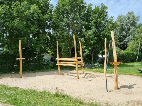 Speeltoestel met houten palen en touwen op een zandondergrond in een groene omgeving van een vakantiepark.
