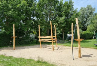 Speeltoestel met houten palen en touwen op een zandondergrond in een groene omgeving van een vakantiepark.