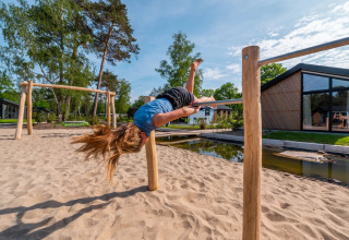 Kind draait op een houten rekstok bij een glamping vakantiepark op een zonnige dag, met zand en huisjes.