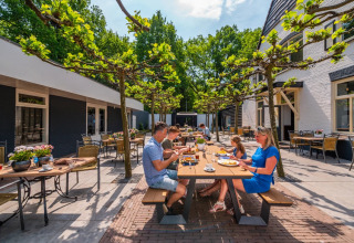 Gezin geniet van een zomerse maaltijd op het terras van een vakantiepark met glamping accommodaties.